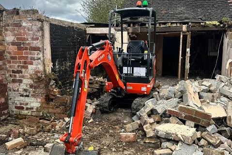A mini digger breaking down an old brick outbuilding, rubble stacked to one side