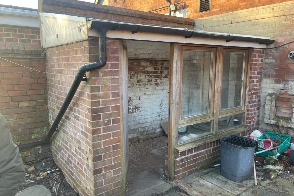 A tired brick lean-to with rotten window frames and a sagging roof, before it comes down