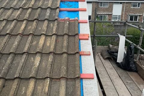 A roof edge stripped back to new battens and membrane, with houses and scaffolding behind